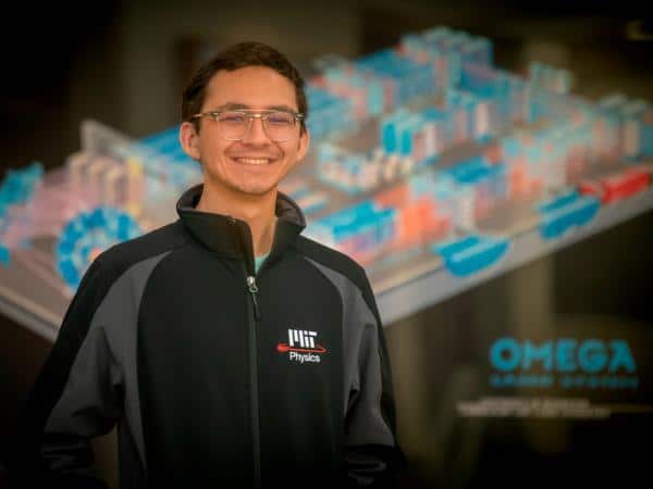 A man in a blue cardigan and glasses smiles at the camera. In the background is a blurry schematic of the OMEGA laser facility.