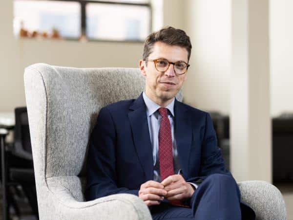 A man in a blue suit and red tie sits in a grey armchair.