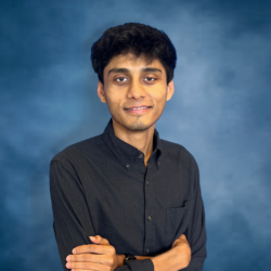 A young brown man with medium-length dark hair smiles shyly while crossing his arms