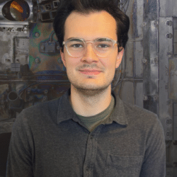 A young white man with coiffed dark hair and glasses gives a slight smile