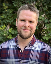 A younger white man with sandy hair and a short sandy beard, wearing a red and navy plaid button down.