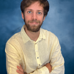 A young white man with medium-lenght light brown hair and bright blue eyes smiles mildly while crossing his arms