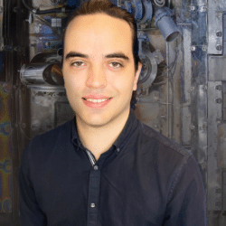 A young white man with dark hair pulled back wears a navy button down and smiles sligtly