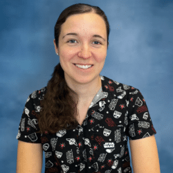 A young white woman with long curly brown hair and a star wars button down gives a friendly smile