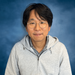 A middle-aged Korean man with medium length black hair and large round glasses gives a small smile