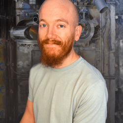 A middle-aged bald white man with a red beard and bright blue eyes smiles mildly