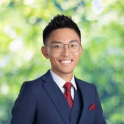 A young Asian man with wire-framed glasses and a big smile wears a navy suit and red tie